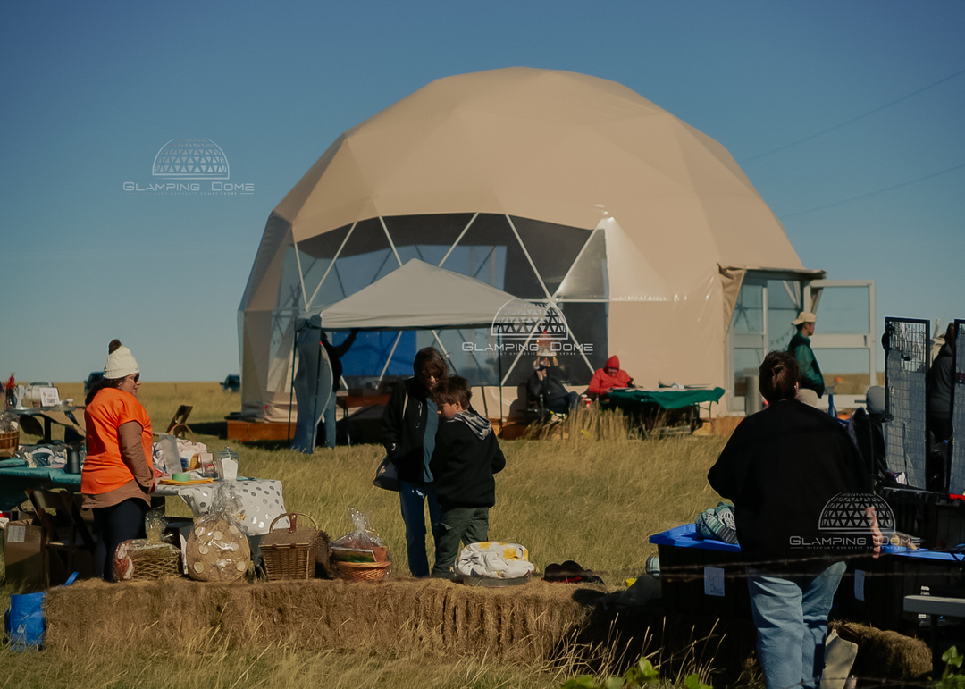 Exterior view of a 32-foot (10-meter) geodesic dome tent, sold by Glamping Dome Store, with a light khaki PVC cover, installed at Healing Grounds by Keys to Hope, Redcliff, Alberta, Canada. The dome is set on a wooden platform and surrounded by market-style activities with vendors and attendees. Transparent panoramic windows provide visibility of the interior, while the scenic prairie landscape and open sky add to the setting's charm, making it ideal for community events and outdoor gatherings.