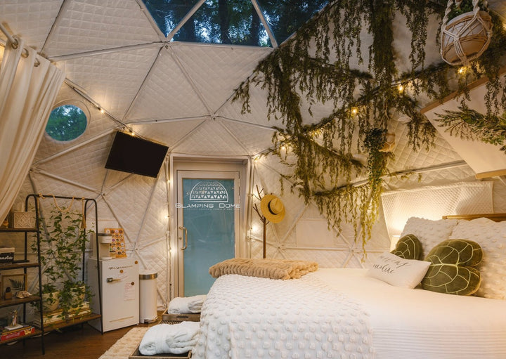 Interior of a 16.4-foot (5-meter) geodesic dome tent at Little Red Homestead, a glamping site in Lake Ann, Michigan, USA, featuring a cozy bedroom setup with plush bed covers, warm lighting, and decorative greenery. The space includes a mini-fridge, shelving unit, and a wall-mounted TV, offering a luxurious and functional glamping experience in a scenic outdoor environment.