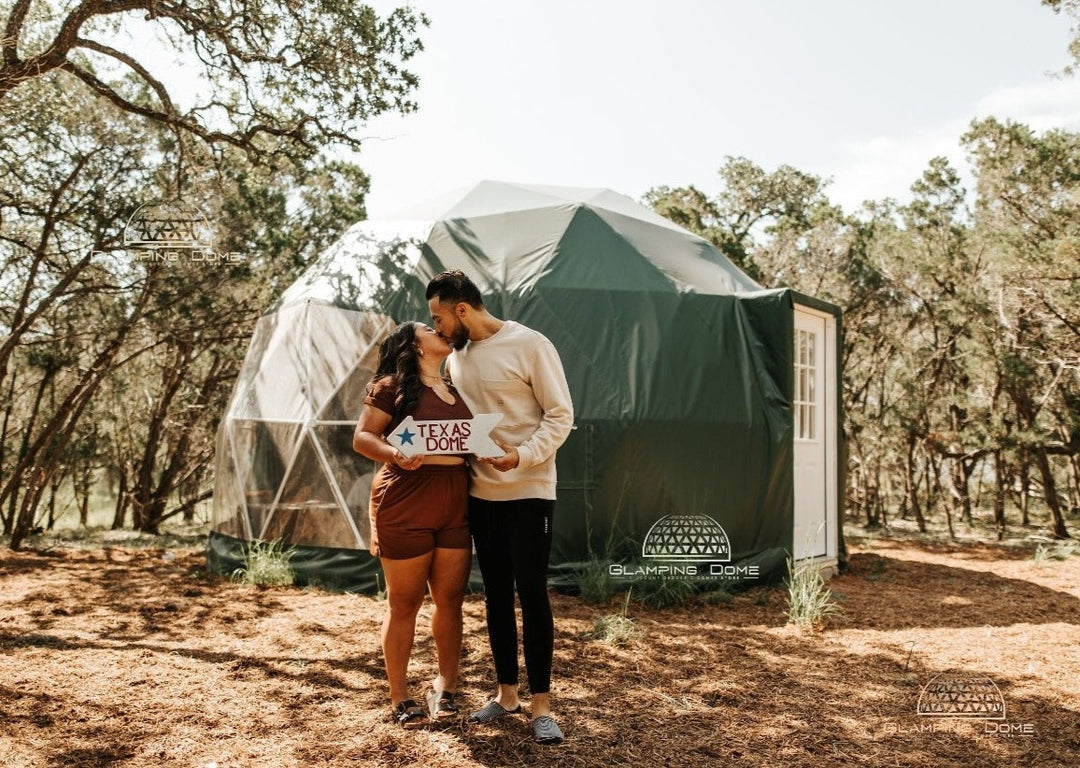 A 19.7-foot (6-meter) geodesic dome tent, sold by Glamping Dome Store, installed at The Cedars Ranch in Wimberley, Texas, USA. The dome features a forest-green weather-resistant PVC cover and a white entrance door. Surrounded by tall cedar trees and natural landscaping, it creates a serene outdoor environment. In the foreground, a couple holds a 'Texas Dome' sign while sharing a kiss, showcasing the dome as a unique and romantic glamping experience.