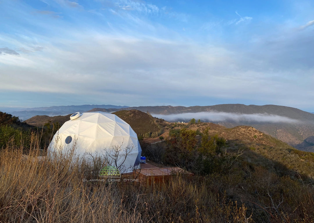 White 16.4-foot (5-meter) geodesic dome tent, sold by Glamping Dome Store, set on a wooden platform in a scenic mountainous area in Westminster, California, USA, surrounded by rolling hills and a misty horizon. Designed for glamping and eco-tourism, the dome blends functionality and aesthetics, offering a unique and unforgettable outdoor living experience.
