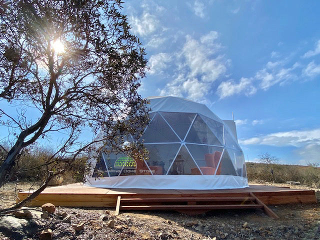 White 16.4-foot (5-meter) geodesic dome tent, sold by Glamping Dome Store, with a transparent PVC panoramic window, set on a wooden platform in a serene desert-like landscape under a clear blue sky in Westminster, California, USA. Perfect for eco-tourism, off-grid living, or luxury camping experiences, showcasing durability and versatility in outdoor environments.
