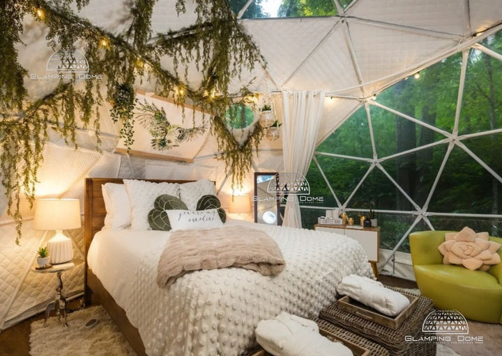 Interior of a 16.4-foot (5-meter) geodesic dome tent at Little Red Homestead, a glamping destination in Lake Ann, Michigan, USA, elegantly furnished with a cozy bed, soft lighting, and decorative greenery. The dome tent features a transparent panoramic window section, providing stunning views of the surrounding forest, ideal for glamping enthusiasts seeking luxury outdoor accommodations.