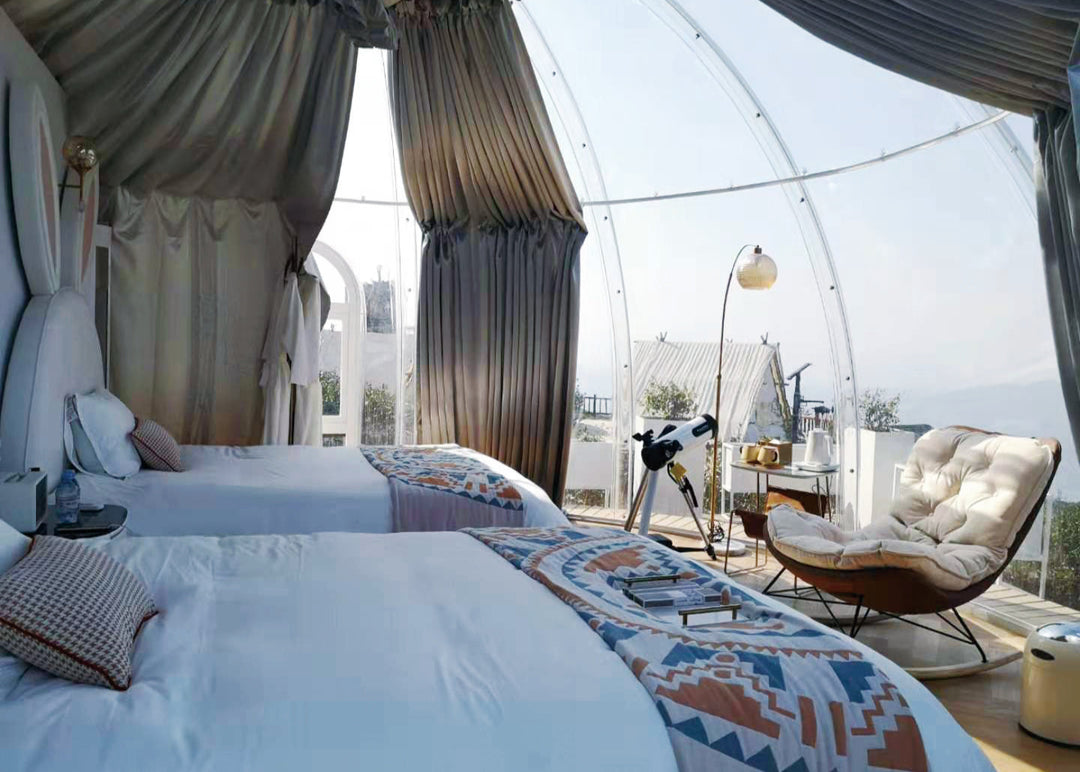 Modern bedroom with two beds, a chair, and large windows with curtains inside a large, hard-shell polycarbonate transparent dome