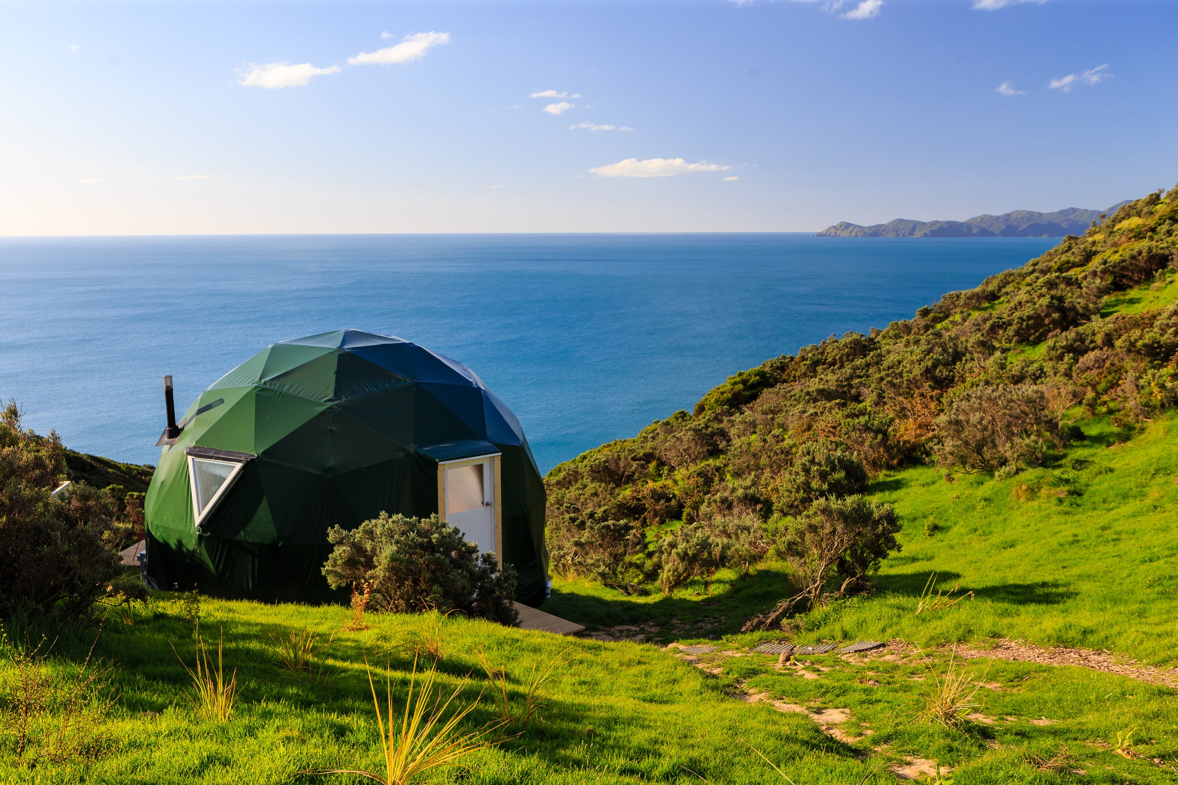 Glamping Dome Store's Geodesic dome on a grassy hill overlooking the Pacific ocean with a clear blue sky of New Zealand.
