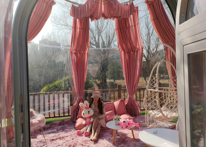 Person sitting on a pink couch with decorative items in a bubble-like structure with pink curtains.