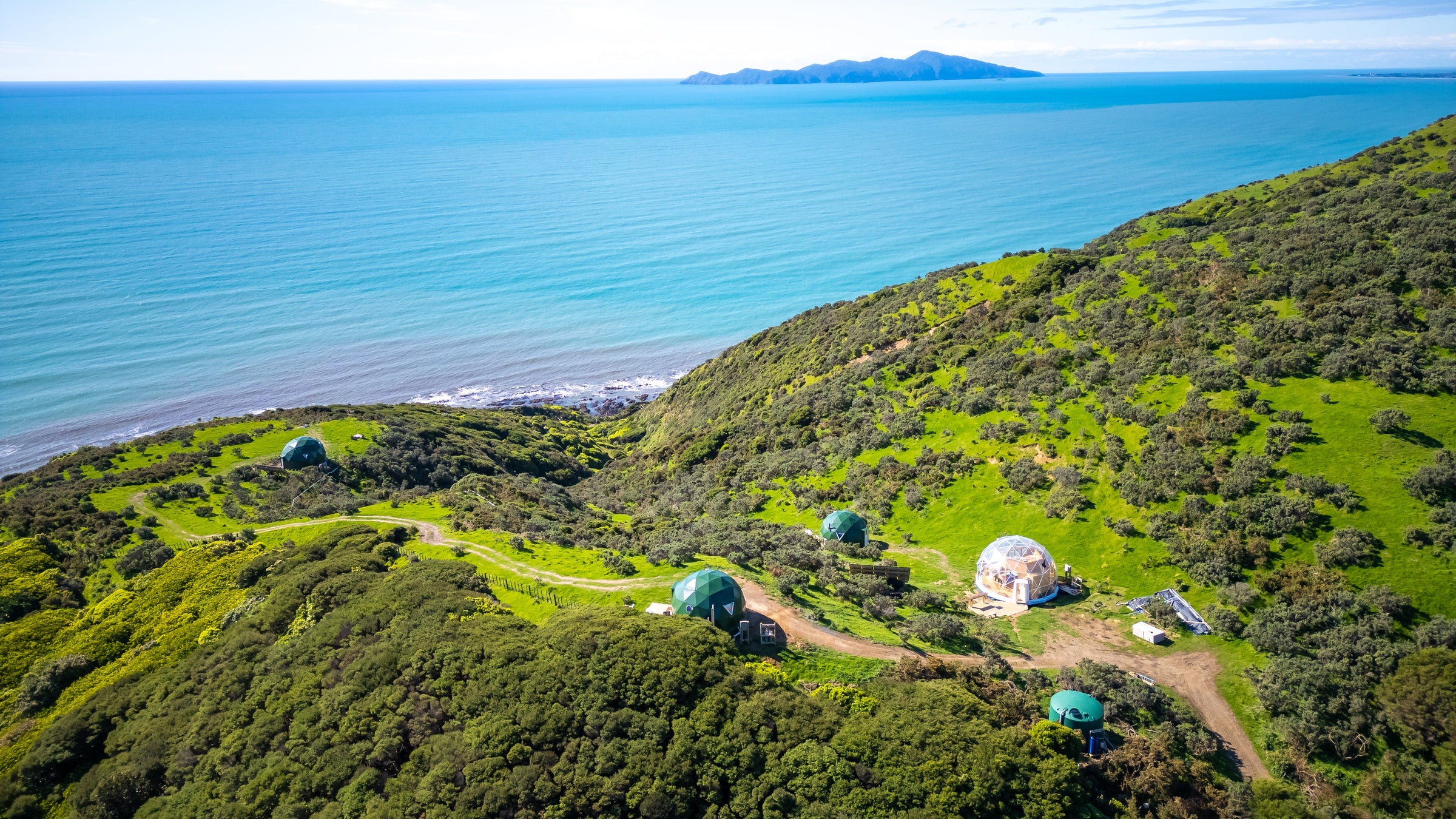 Houses on a hill overlooking a vast blue Pacific ocean with clear skies of the New Zealand seaside. Project domes were supplied by Glamping Dome Store.