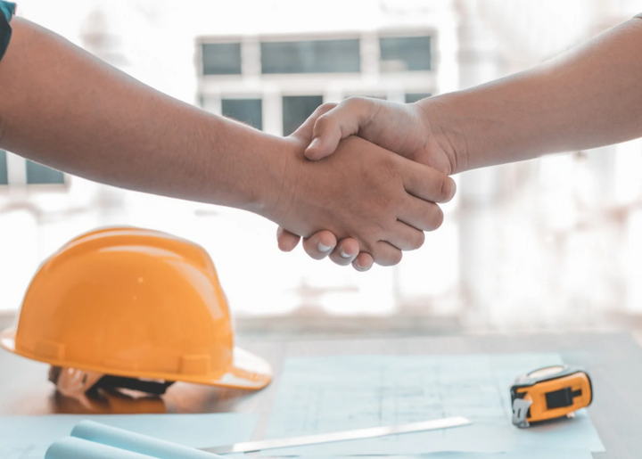 Two professionals shaking hands over architectural blueprints, symbolizing trust and reliability in structural engineering. A yellow hard hat and measuring tools on the table reinforce a commitment to safety and precision. This image represents Glamping Dome Store’s dedication to providing professionally engineered reports, ensuring geodesic dome tents meet wind and snow load requirements for secure installations.