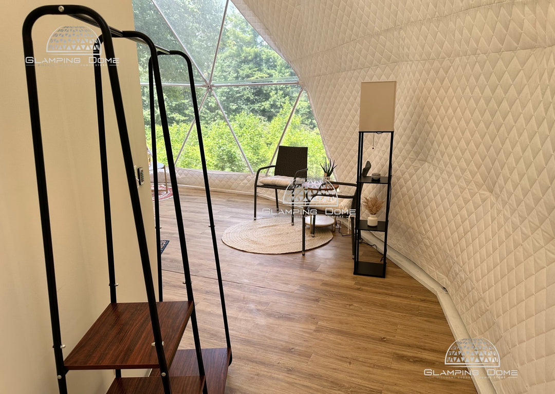 Interior of a 29.5-foot (9-meter) geodesic dome tent by Glamping Dome Store, located at Big Little Clouds in Charlotte, North Carolina, USA. The dome features a quilted synthetic insulation fabric layer, available in many colors, along with modern furnishings, a seating area with a round rug, and large panoramic window offering a view of lush greenery outside.