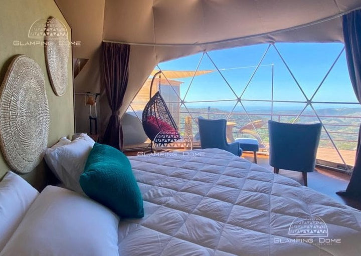 Interior of a geodesic dome tent kit sold by Glamping Dome Store, featuring a luxurious bed with crisp white linens and teal accent pillows. The cozy space is adorned with woven wall art and floor-to-ceiling curtains framing a panoramic view through the transparent PVC windows. Outside, a hanging egg chair and seating area on the wooden deck provide a perfect spot to relax and enjoy scenic vistas. This setup highlights the elegance and versatility of glamping dome tents.
