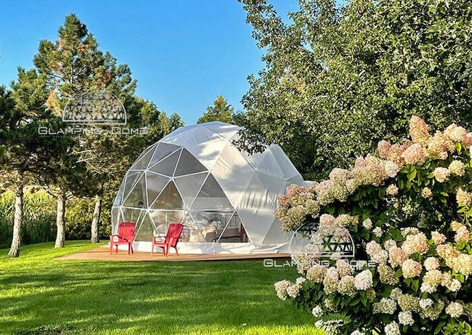 23-foot (7-meter) geodesic dome tent, a project by Glamping Dome Store, located at Maison des Prés with Dome & Jacuzzi in Saint-Basile-le-Grand, Quebec, Canada. The dome features a transparent extended PVC panoramic window, a weather-resistant PVC white cover, and is set on a wooden platform surrounded by lush greenery and blooming hydrangeas. Red Adirondack chairs add a cozy outdoor touch, creating an inviting space for glamping and relaxation.