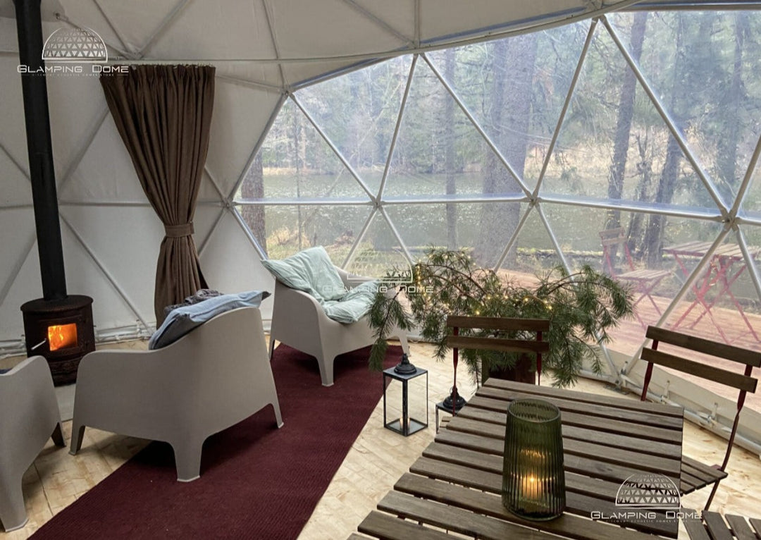 Interior view of a 23-foot (8 m) geodesic dome tent, sold by Glamping Dome Store, set in a serene forest location. The cozy setup includes modern chairs, a small wood-burning stove, a dining table with a lantern, and festive greenery. The large panoramic window provides stunning views of the surrounding trees and a tranquil lake. This inviting space highlights the Glamping Dome Store's expertise in providing dome tent structures and accessories, ideal for relaxing retreats or unique outdoor accommodations.