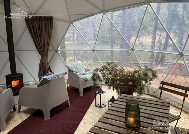 Interior view of a 23-foot (8 m) geodesic dome tent, sold by Glamping Dome Store, set in a serene forest location. The cozy setup includes modern chairs, a small wood-burning stove, a dining table with a lantern, and festive greenery. The large panoramic window provides stunning views of the surrounding trees and a tranquil lake. This inviting space highlights the Glamping Dome Store's expertise in providing dome tent structures and accessories, ideal for relaxing retreats or unique outdoor accommodations.