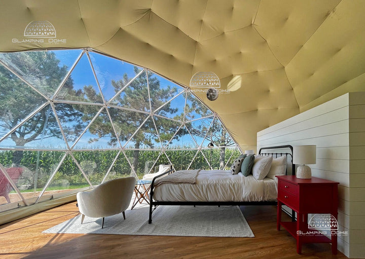 Interior of a 23-foot (7-meter) geodesic dome tent kit, a project by Glamping Dome Store, located at Maison des Prés with Dome & Jacuzzi in Saint-Basile-le-Grand, Quebec, Canada. The space features a luxurious setup with a cozy bed, plush bedding, a bright red nightstand, and a comfortable armchair. The transparent PVC panoramic window offers stunning views of the surrounding greenery and a vibrant blue sky, creating a perfect glamping retreat.